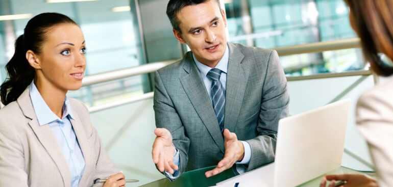 Professional business consultants discussing strategy with a client in a modern office, reviewing charts and data on a laptop during a meeting.